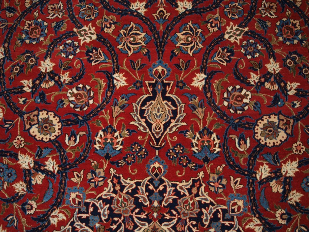 A large hand-knotted Persian Isfahan area rug featuring a red field filled with intricate floral arabesque vines and a central medallion surrounded by a dark blue ornate border. View X