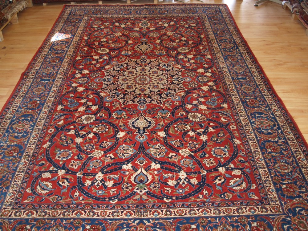A large hand-knotted Persian Isfahan area rug featuring a red field filled with intricate floral arabesque vines and a central medallion surrounded by a dark blue ornate border. View XI