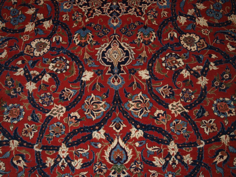 A large hand-knotted Persian Isfahan area rug featuring a red field filled with intricate floral arabesque vines and a central medallion surrounded by a dark blue ornate border. View XII
