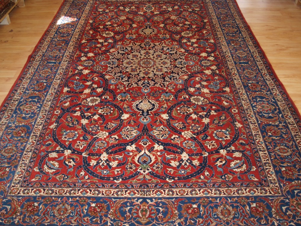 A large hand-knotted Persian Isfahan area rug featuring a red field filled with intricate floral arabesque vines and a central medallion surrounded by a dark blue ornate border. View XIII