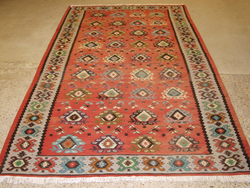 A handwoven flatweave kilim rug featuring a terracotta red central field with repeating multi-coloured geometric motifs and a decorative outer border.