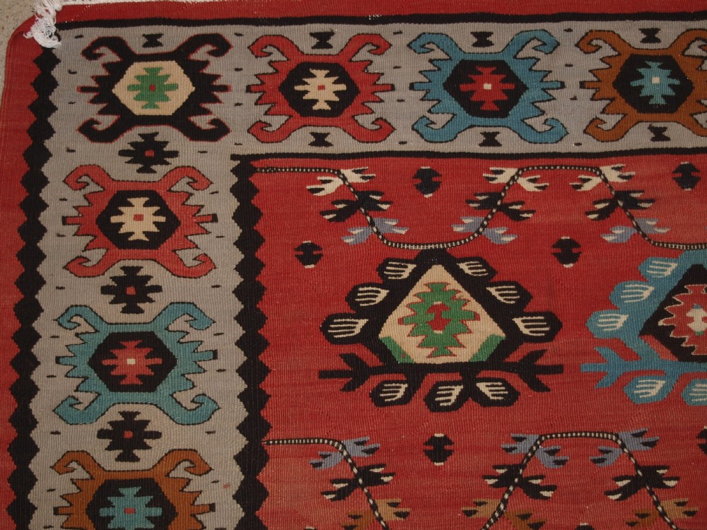 A handwoven flatweave kilim rug featuring a terracotta red central field with repeating multi-coloured geometric motifs and a decorative outer border. View II