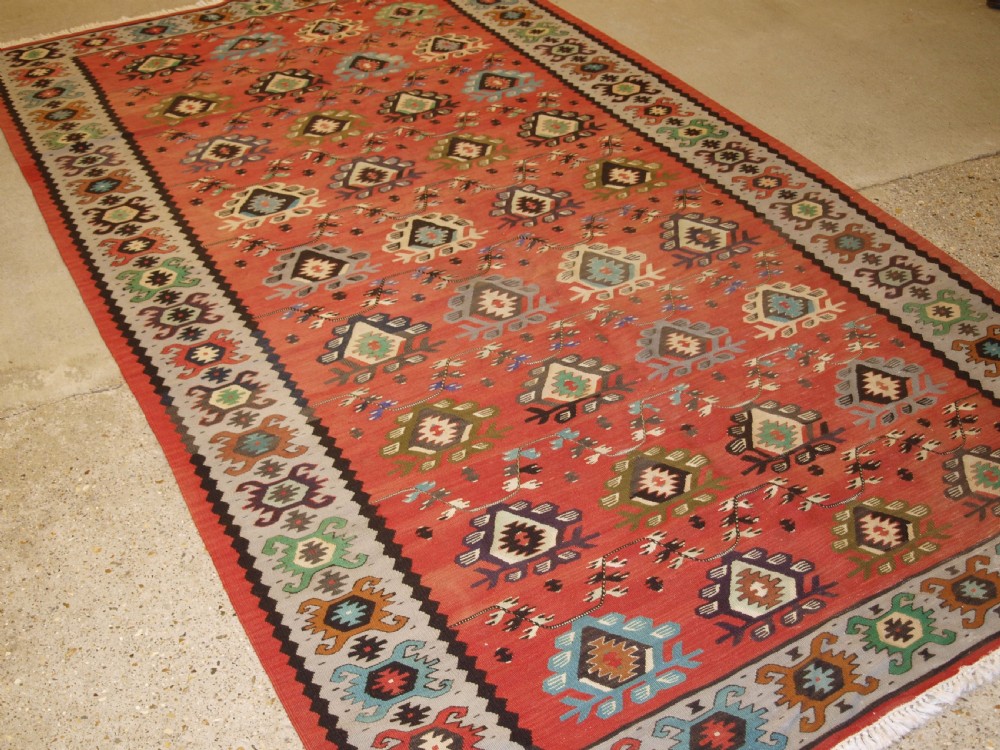 A handwoven flatweave kilim rug featuring a terracotta red central field with repeating multi-coloured geometric motifs and a decorative outer border. View III