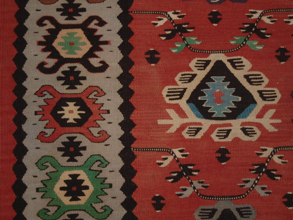 A handwoven flatweave kilim rug featuring a terracotta red central field with repeating multi-coloured geometric motifs and a decorative outer border. View IV