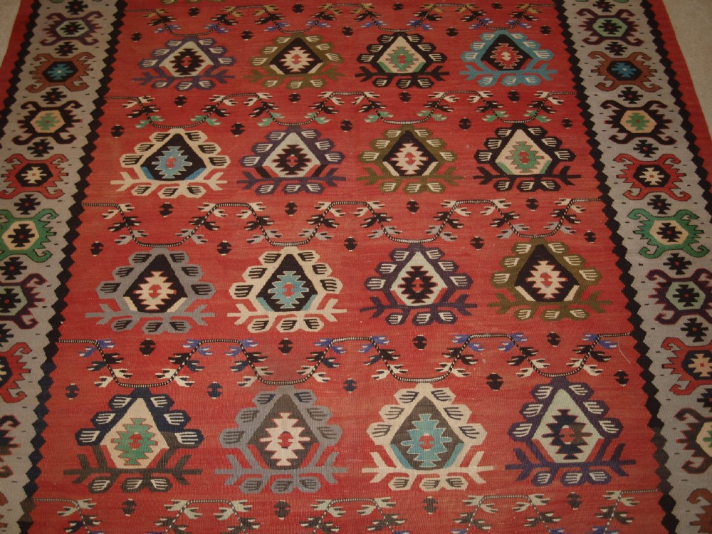 A handwoven flatweave kilim rug featuring a terracotta red central field with repeating multi-coloured geometric motifs and a decorative outer border. View V
