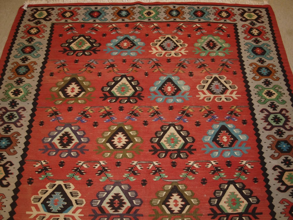 A handwoven flatweave kilim rug featuring a terracotta red central field with repeating multi-coloured geometric motifs and a decorative outer border. View VI