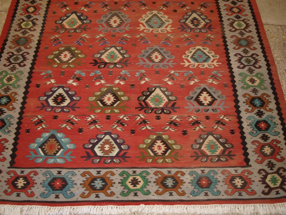 A handwoven flatweave kilim rug featuring a terracotta red central field with repeating multi-coloured geometric motifs and a decorative outer border. View VII