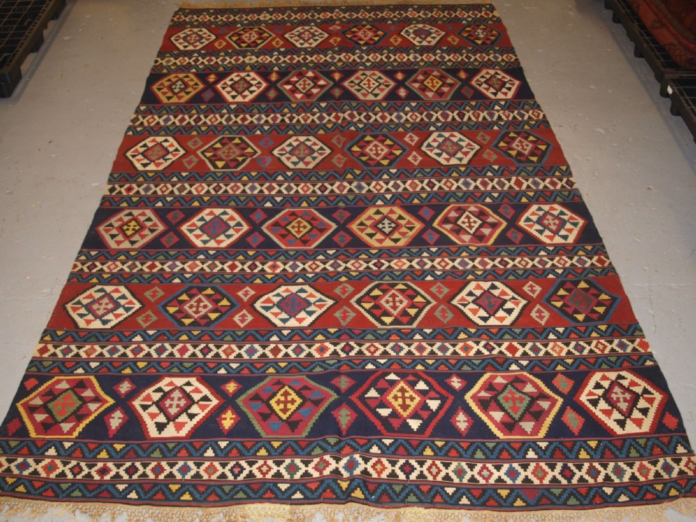A large flat-woven antique Caucasian Shirvan kilim rug featuring horizontal bands of geometric diamond motifs in red, navy blue, green, and yellow.