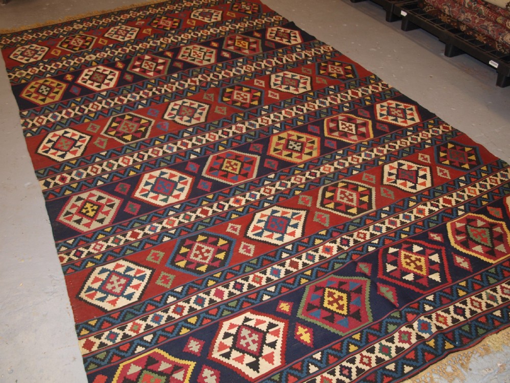 A large flat-woven antique Caucasian Shirvan kilim rug featuring horizontal bands of geometric diamond motifs in red, navy blue, green, and yellow. View X