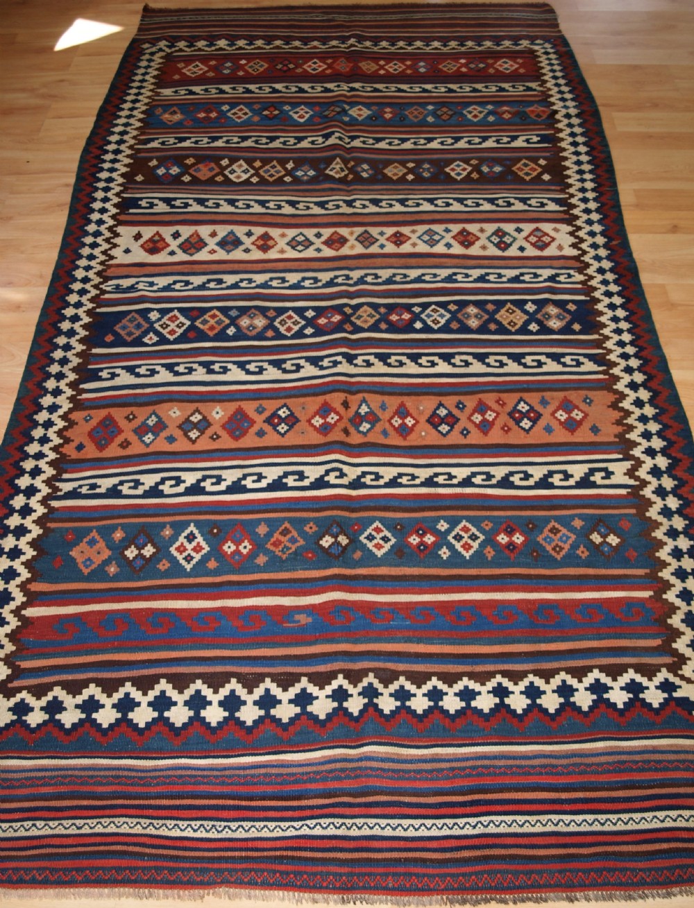 An antique handwoven Persian Varamin kilim rug featuring horizontal bands of blue, terracotta, and cream with repeating geometric diamond motifs.