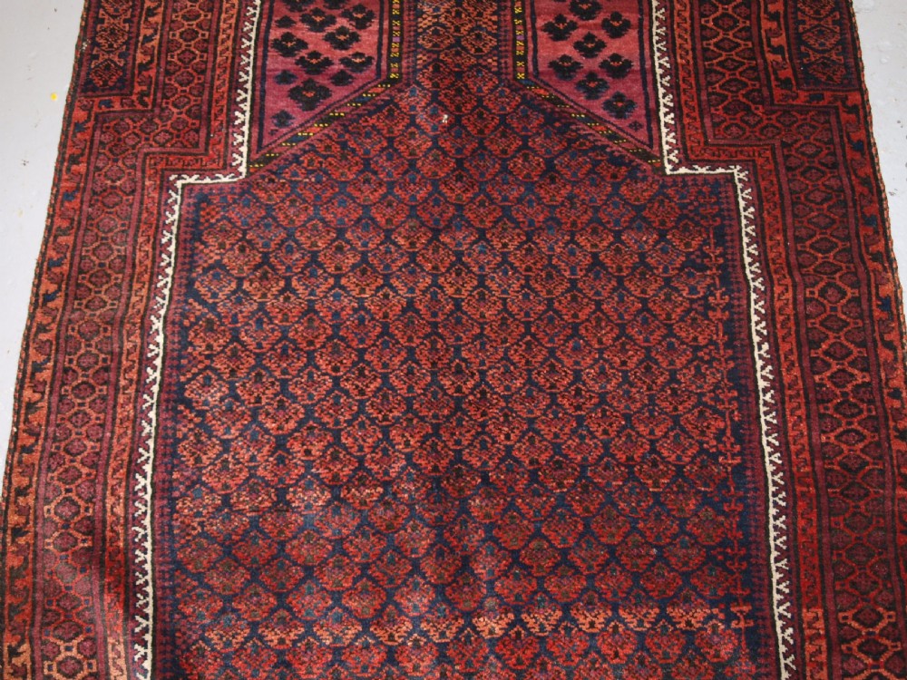 An antique hand-knotted Baluch prayer rug featuring a pointed mihrab arch motif, geometric lattice patterning, and a dark red and indigo colour palette. View VI