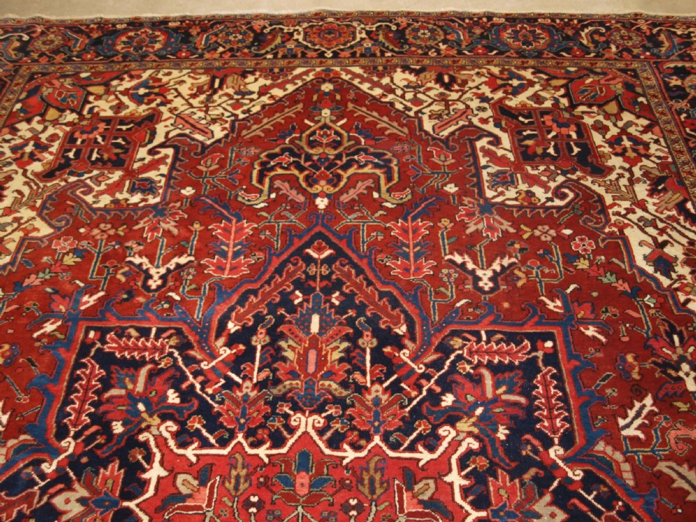 A large hand-knotted Persian Heriz carpet featuring a prominent geometric central medallion in dark navy and red, framed by cream spandrels and a wide dark border. View IV
