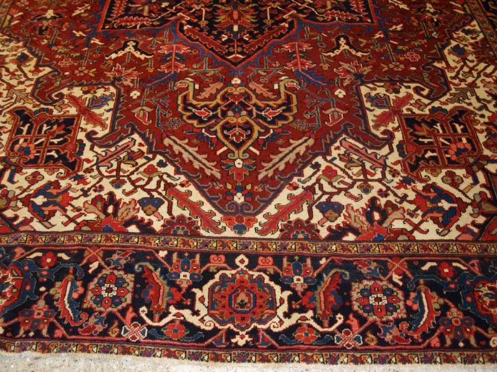 A large hand-knotted Persian Heriz carpet featuring a prominent geometric central medallion in dark navy and red, framed by cream spandrels and a wide dark border. View VI