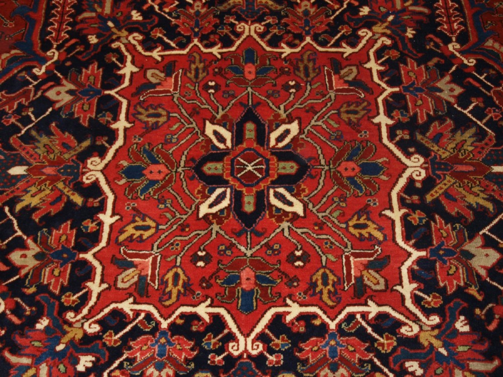 A large hand-knotted Persian Heriz carpet featuring a prominent geometric central medallion in dark navy and red, framed by cream spandrels and a wide dark border. View VII