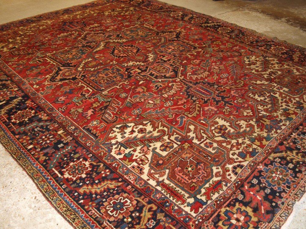 An antique Persian Heriz wool carpet featuring a bold red field with a central geometric medallion, ivory spandrels, and navy blue floral borders.