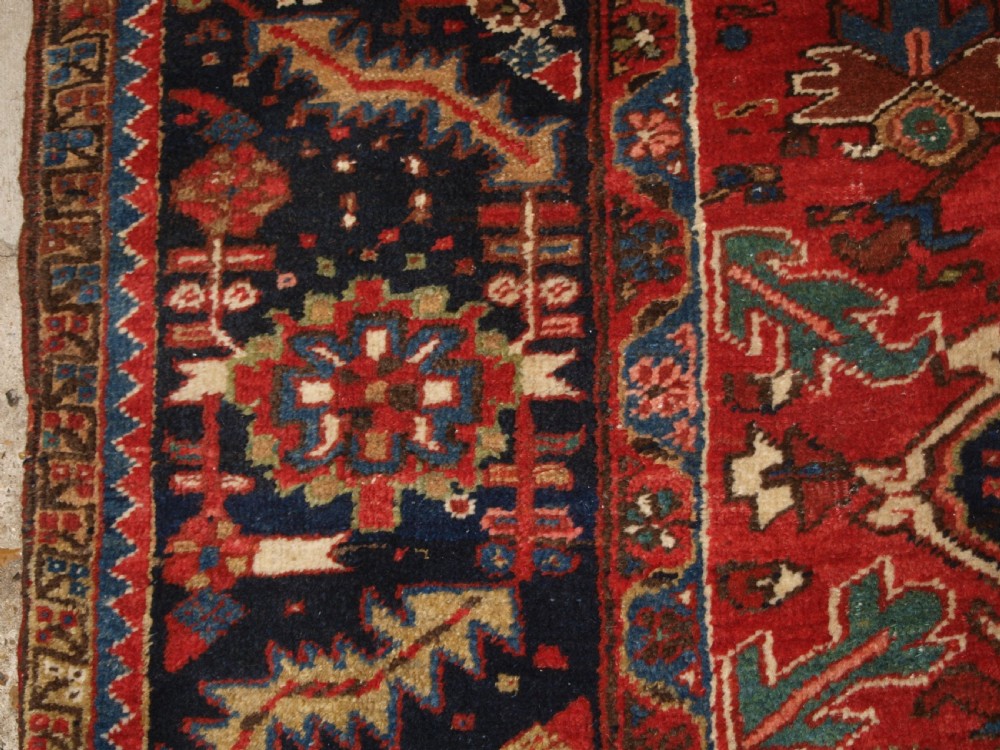 An antique Persian Heriz wool carpet featuring a bold red field with a central geometric medallion, ivory spandrels, and navy blue floral borders. View III