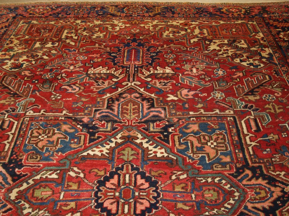 An antique Persian Heriz wool carpet featuring a bold red field with a central geometric medallion, ivory spandrels, and navy blue floral borders. View IV