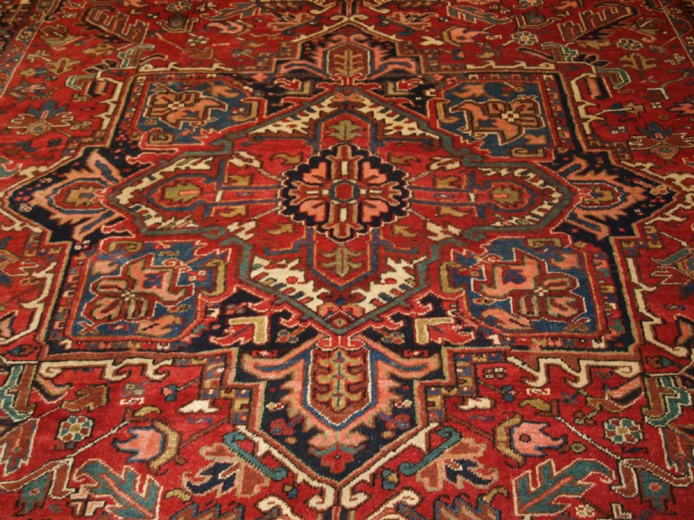 An antique Persian Heriz wool carpet featuring a bold red field with a central geometric medallion, ivory spandrels, and navy blue floral borders. View V