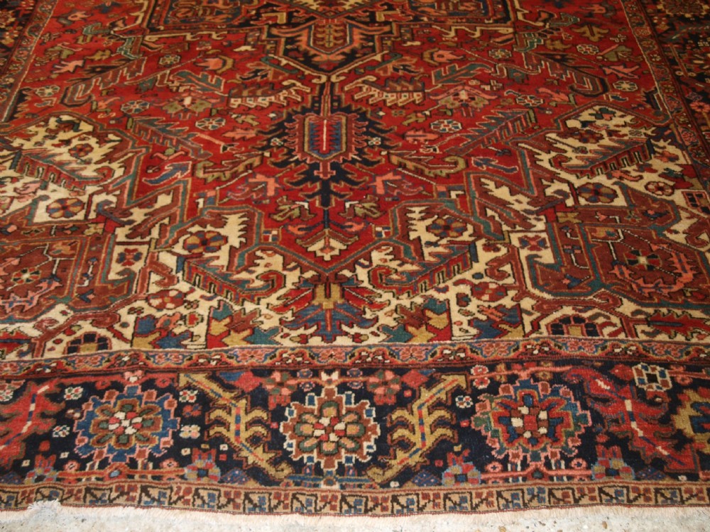 An antique Persian Heriz wool carpet featuring a bold red field with a central geometric medallion, ivory spandrels, and navy blue floral borders. View VI