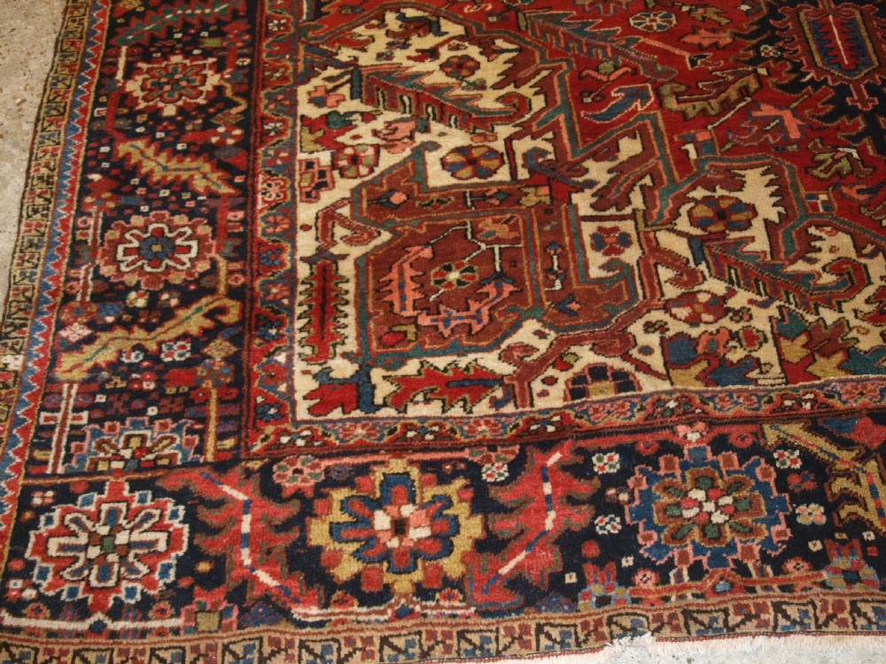 An antique Persian Heriz wool carpet featuring a bold red field with a central geometric medallion, ivory spandrels, and navy blue floral borders. View VII
