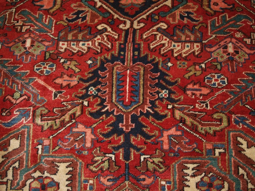 An antique Persian Heriz wool carpet featuring a bold red field with a central geometric medallion, ivory spandrels, and navy blue floral borders. View VIII