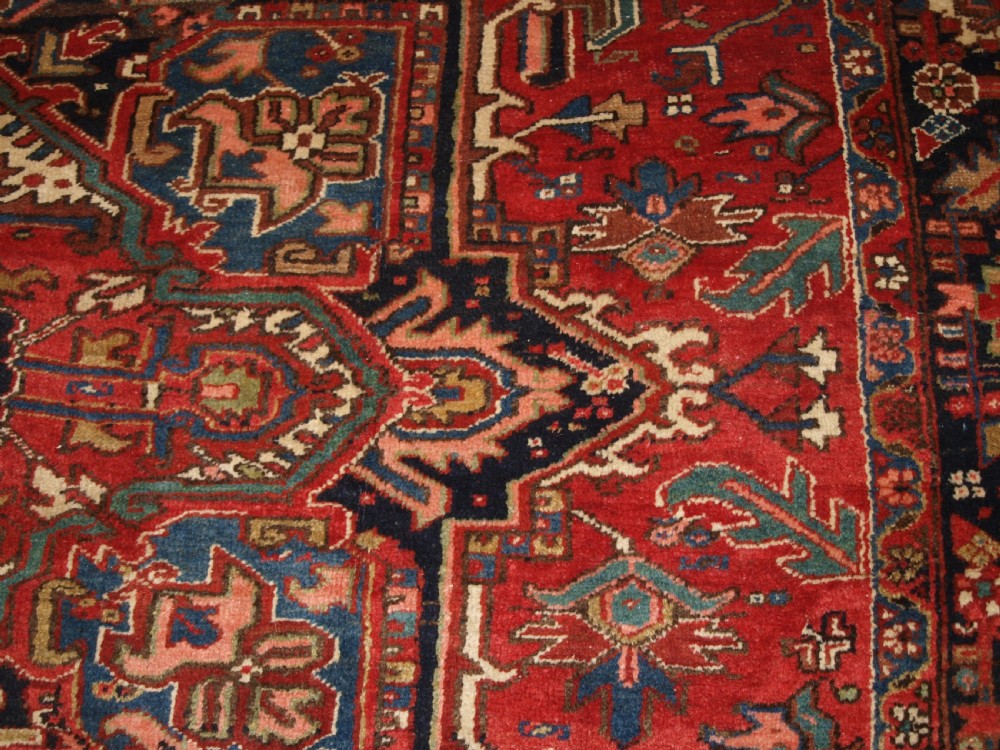 An antique Persian Heriz wool carpet featuring a bold red field with a central geometric medallion, ivory spandrels, and navy blue floral borders. View IX
