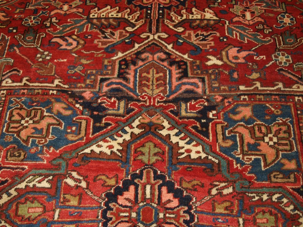 An antique Persian Heriz wool carpet featuring a bold red field with a central geometric medallion, ivory spandrels, and navy blue floral borders. View X
