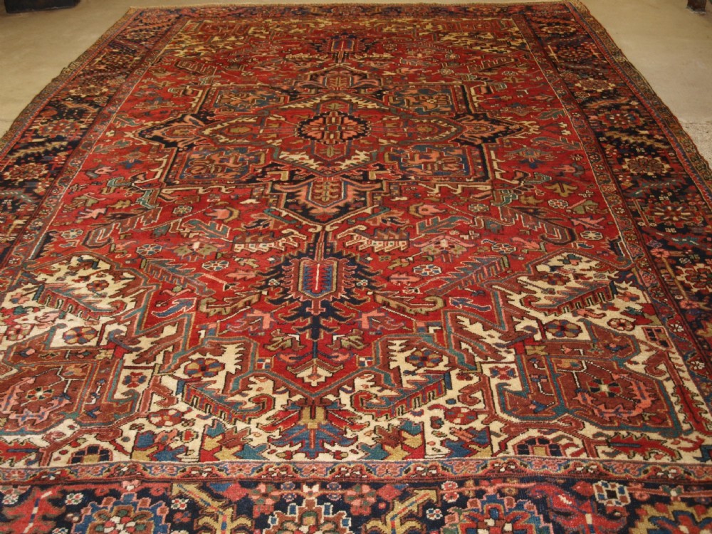 An antique Persian Heriz wool carpet featuring a bold red field with a central geometric medallion, ivory spandrels, and navy blue floral borders. View XI