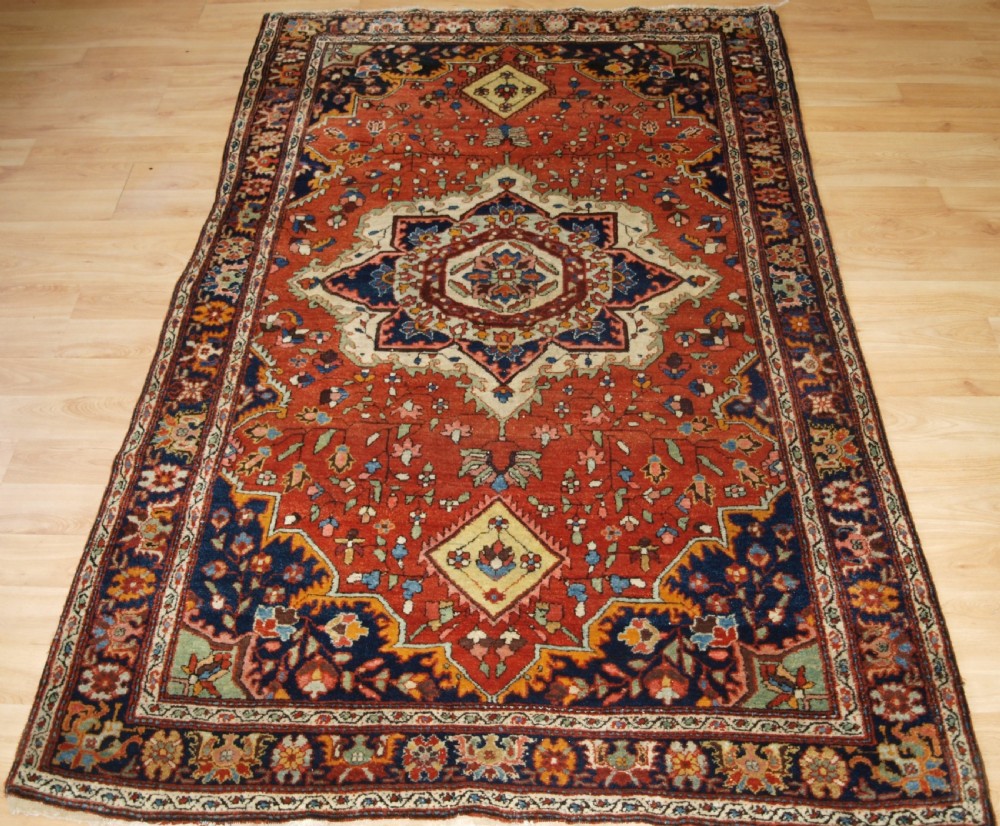 A hand-knotted antique Persian Sarouk wool rug with a prominent central star medallion, floral motifs, terracotta red field, and navy blue border.