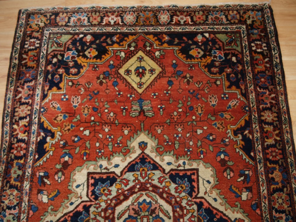 A hand-knotted antique Persian Sarouk wool rug with a prominent central star medallion, floral motifs, terracotta red field, and navy blue border. View V