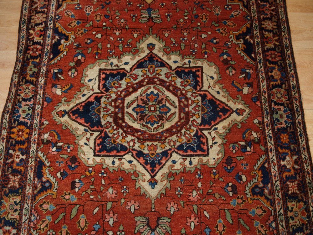 A hand-knotted antique Persian Sarouk wool rug with a prominent central star medallion, floral motifs, terracotta red field, and navy blue border. View VI