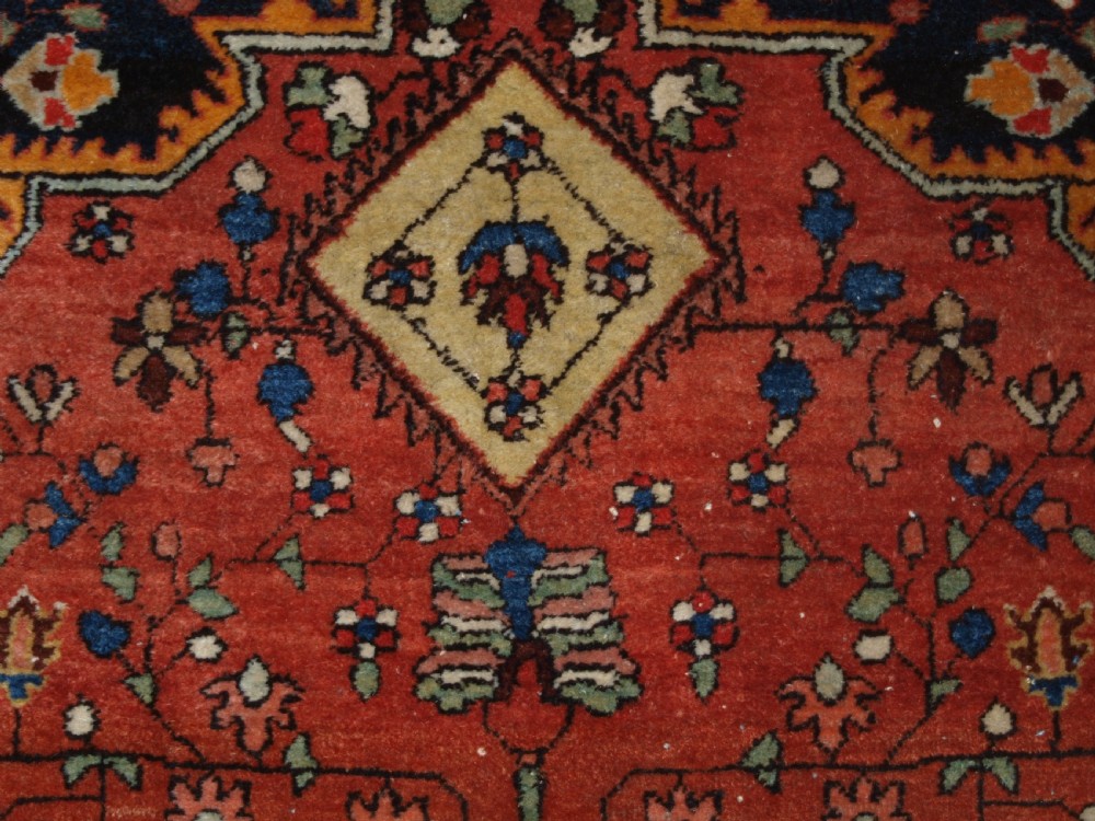 A hand-knotted antique Persian Sarouk wool rug with a prominent central star medallion, floral motifs, terracotta red field, and navy blue border. View VII