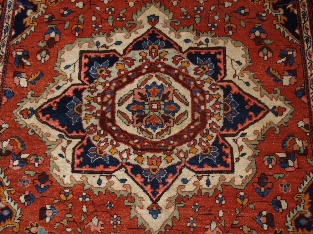 A hand-knotted antique Persian Sarouk wool rug with a prominent central star medallion, floral motifs, terracotta red field, and navy blue border. View IX