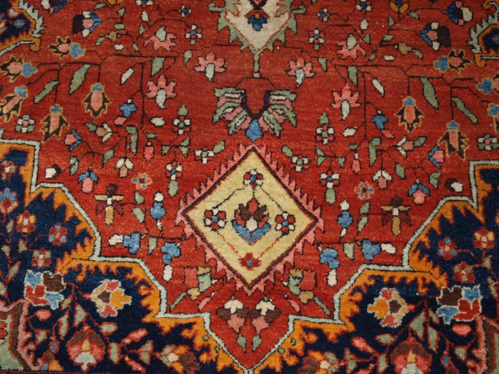 A hand-knotted antique Persian Sarouk wool rug with a prominent central star medallion, floral motifs, terracotta red field, and navy blue border. View X