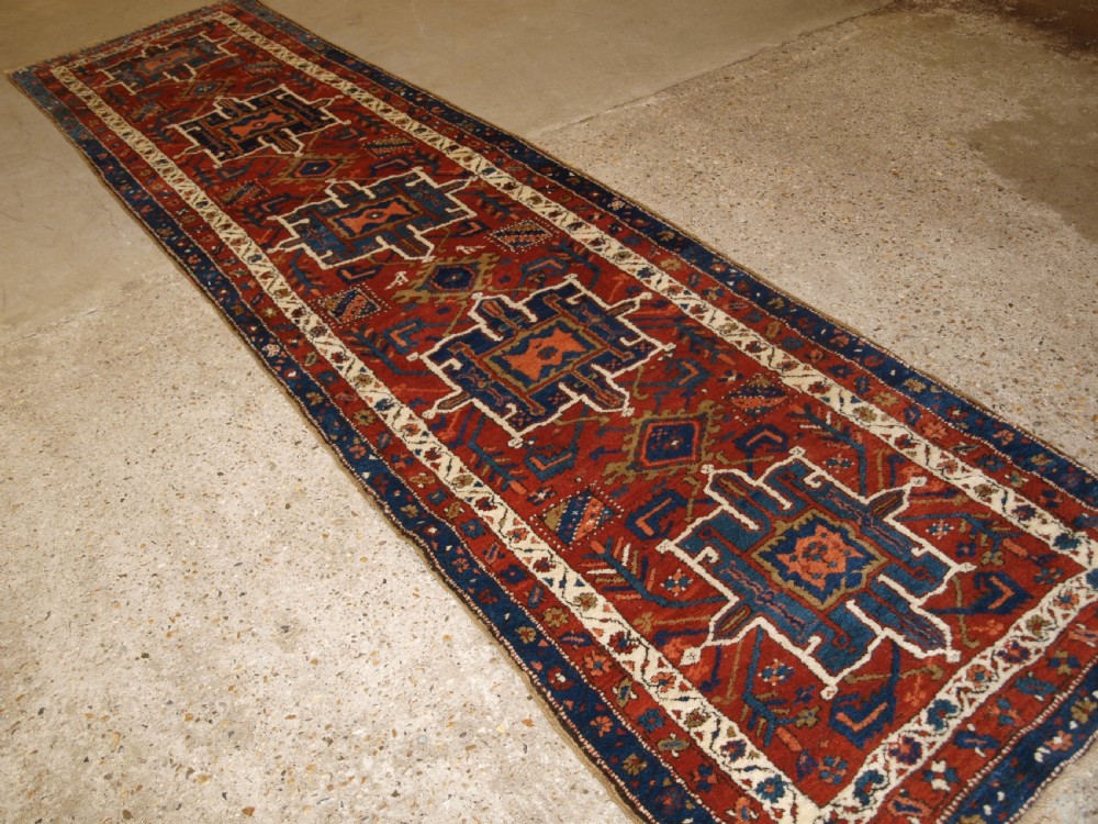 An antique Persian Karaja long runner rug featuring geometric hooked medallions on a terracotta red field framed by ivory and navy blue decorative borders. View II