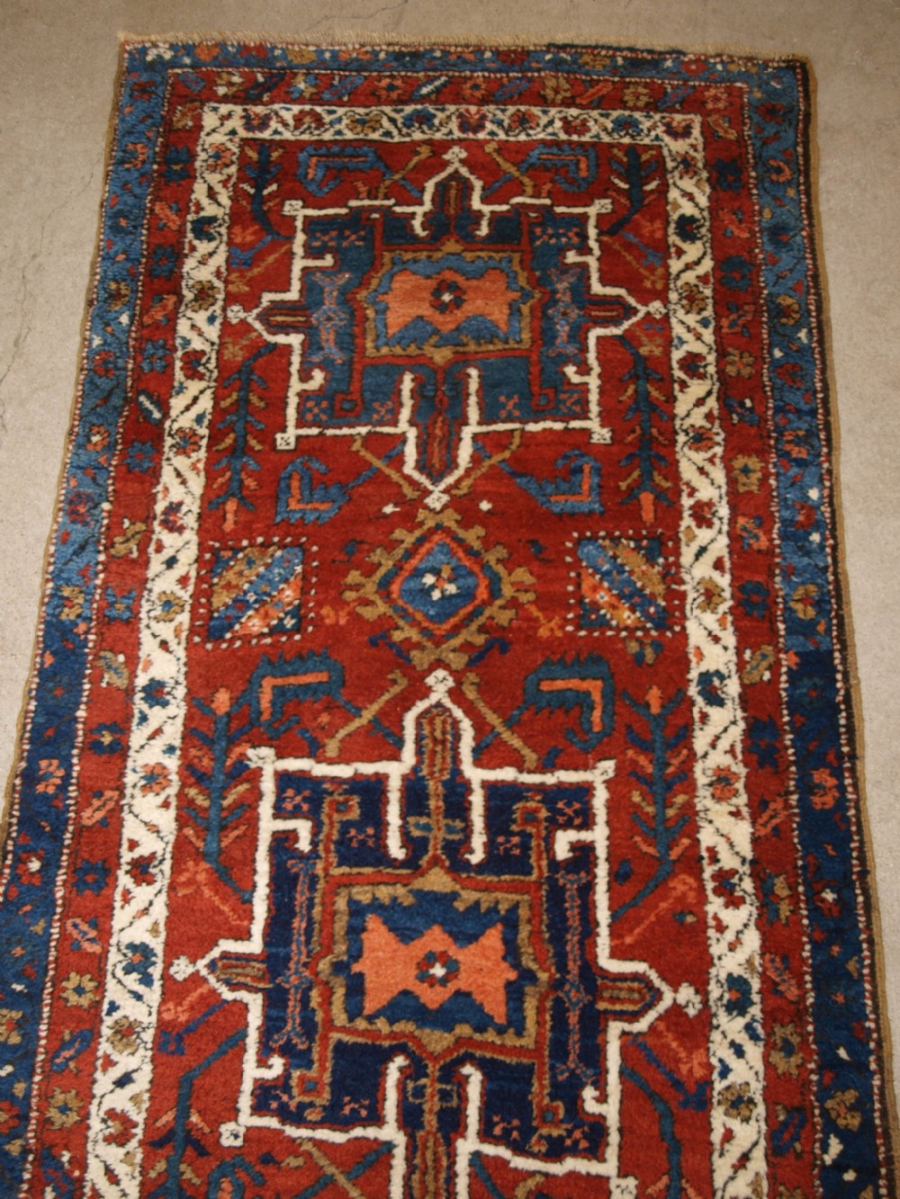 An antique Persian Karaja long runner rug featuring geometric hooked medallions on a terracotta red field framed by ivory and navy blue decorative borders. View V