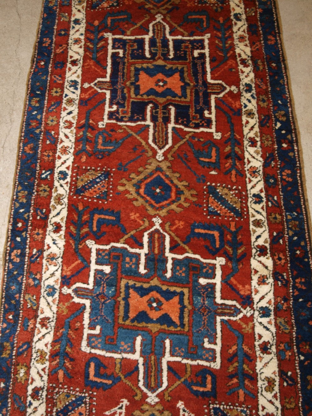 An antique Persian Karaja long runner rug featuring geometric hooked medallions on a terracotta red field framed by ivory and navy blue decorative borders. View VI