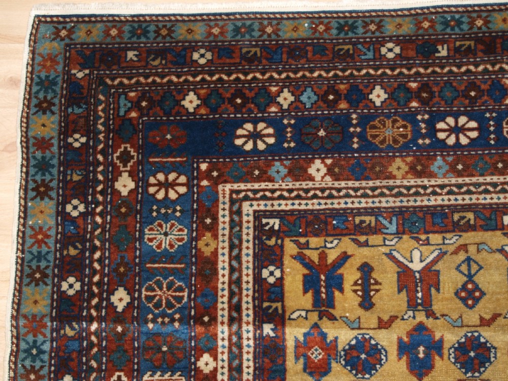 A hand-knotted wool rug featuring a central yellow field decorated with geometric motifs, enclosed by multi-tiered navy blue and terracotta patterned borders. View III