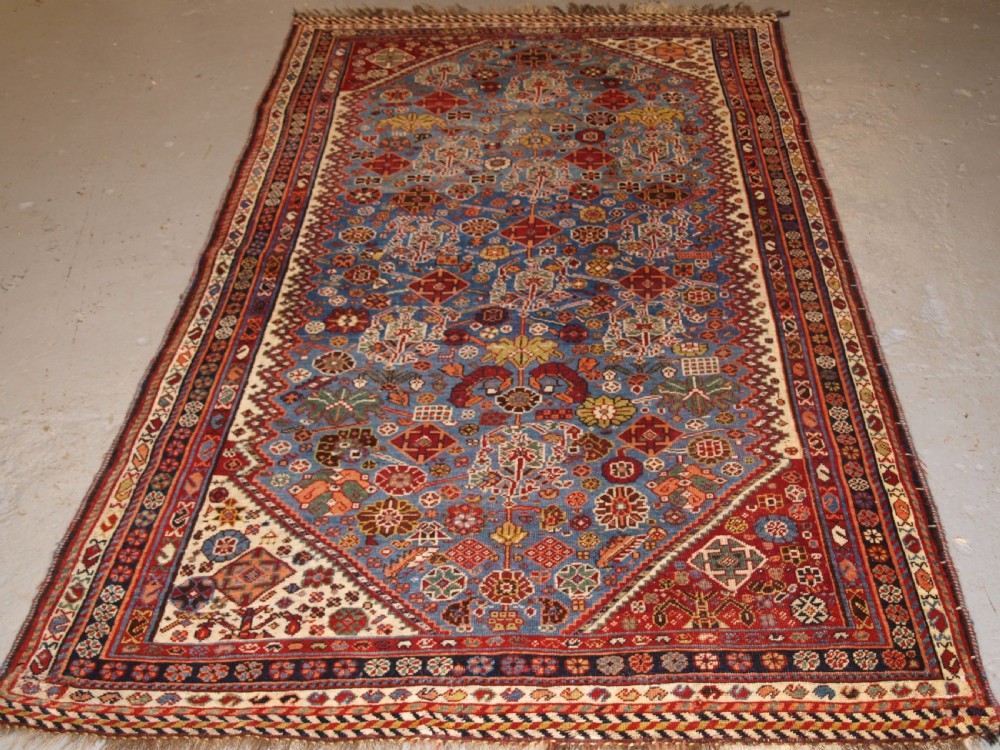 An antique hand-knotted Qashqai Persian rug featuring a central blue field filled with red, white, and yellow geometric tribal motifs framed by a multi-stripe border.