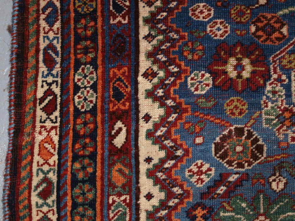 An antique hand-knotted Qashqai Persian rug featuring a central blue field filled with red, white, and yellow geometric tribal motifs framed by a multi-stripe border. View IV