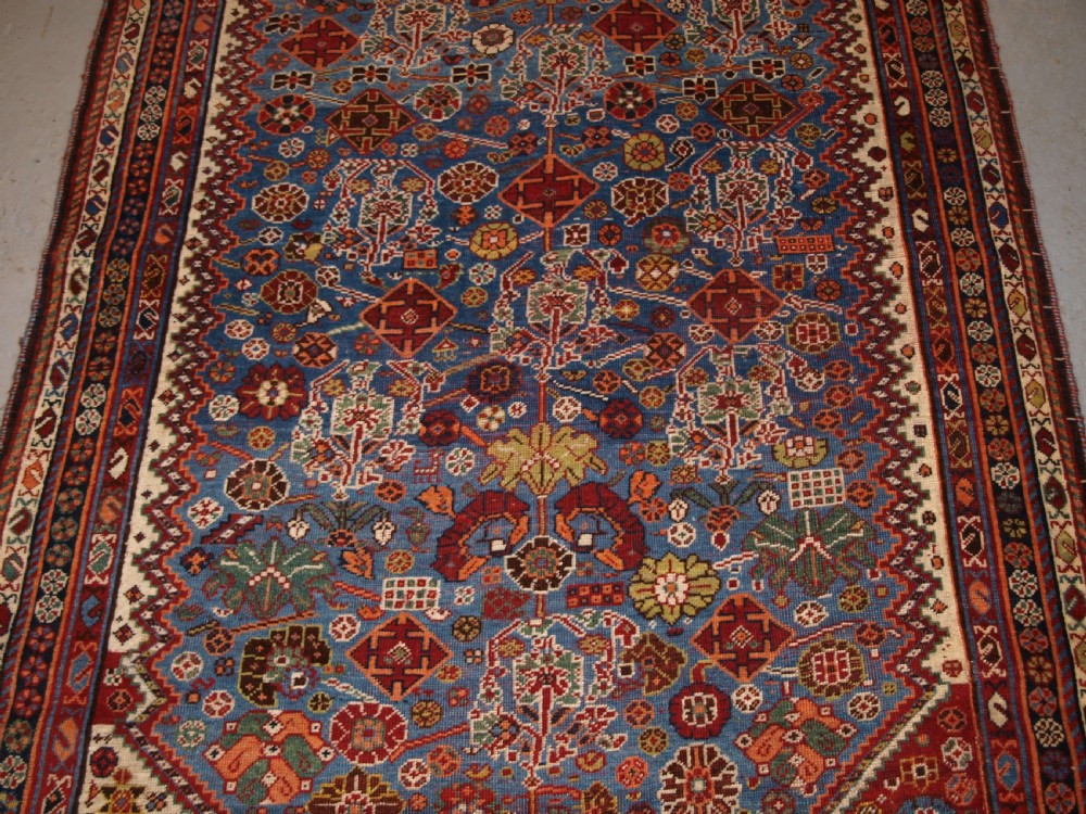 An antique hand-knotted Qashqai Persian rug featuring a central blue field filled with red, white, and yellow geometric tribal motifs framed by a multi-stripe border. View VI