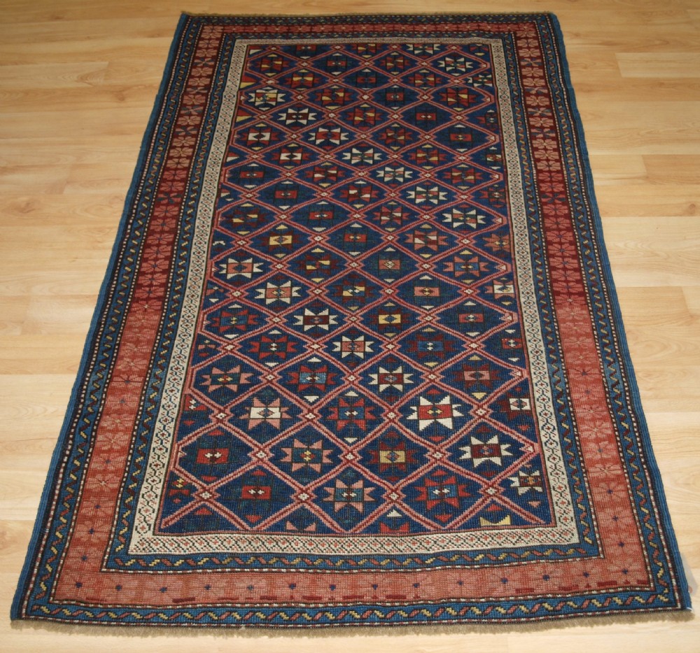 An antique hand-knotted Caucasian rug featuring a central navy blue field with a geometric star-and-lattice pattern framed by a reddish-pink patterned border.