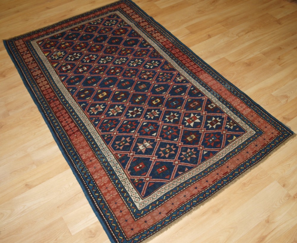 An antique hand-knotted Caucasian rug featuring a central navy blue field with a geometric star-and-lattice pattern framed by a reddish-pink patterned border. View II