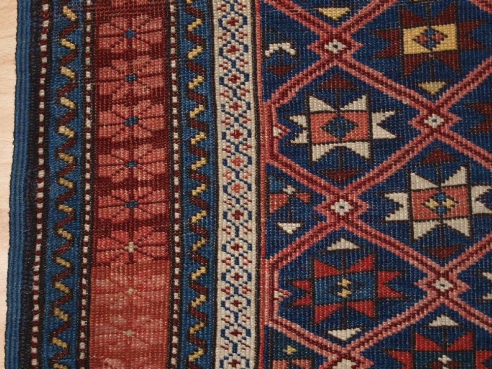 An antique hand-knotted Caucasian rug featuring a central navy blue field with a geometric star-and-lattice pattern framed by a reddish-pink patterned border. View IV