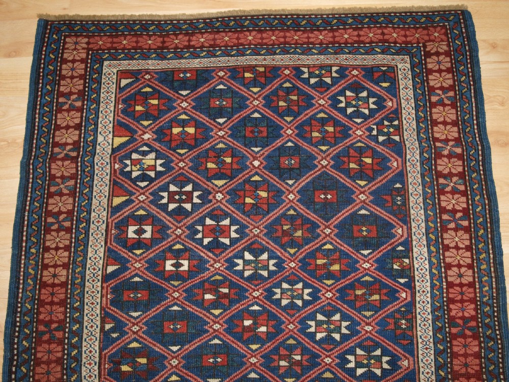 An antique hand-knotted Caucasian rug featuring a central navy blue field with a geometric star-and-lattice pattern framed by a reddish-pink patterned border. View V