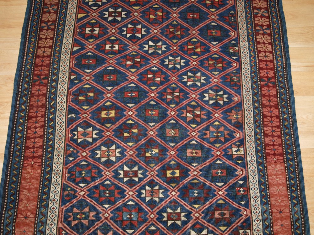 An antique hand-knotted Caucasian rug featuring a central navy blue field with a geometric star-and-lattice pattern framed by a reddish-pink patterned border. View VI