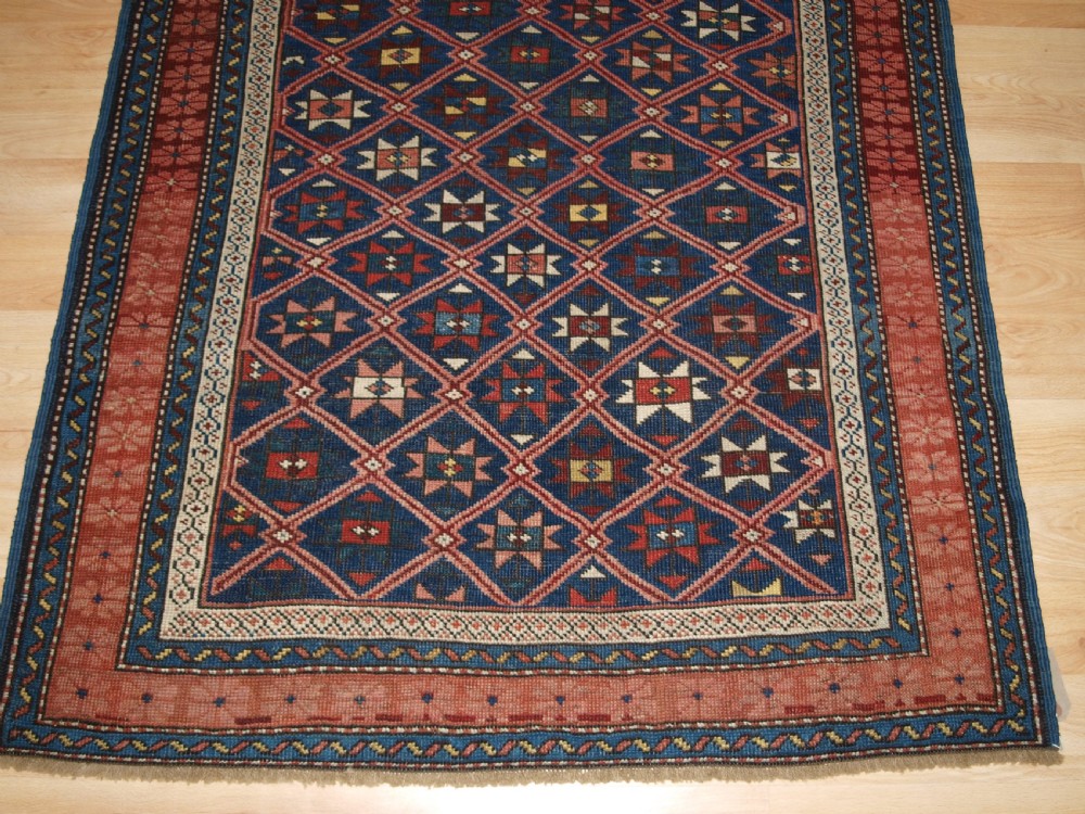 An antique hand-knotted Caucasian rug featuring a central navy blue field with a geometric star-and-lattice pattern framed by a reddish-pink patterned border. View VII