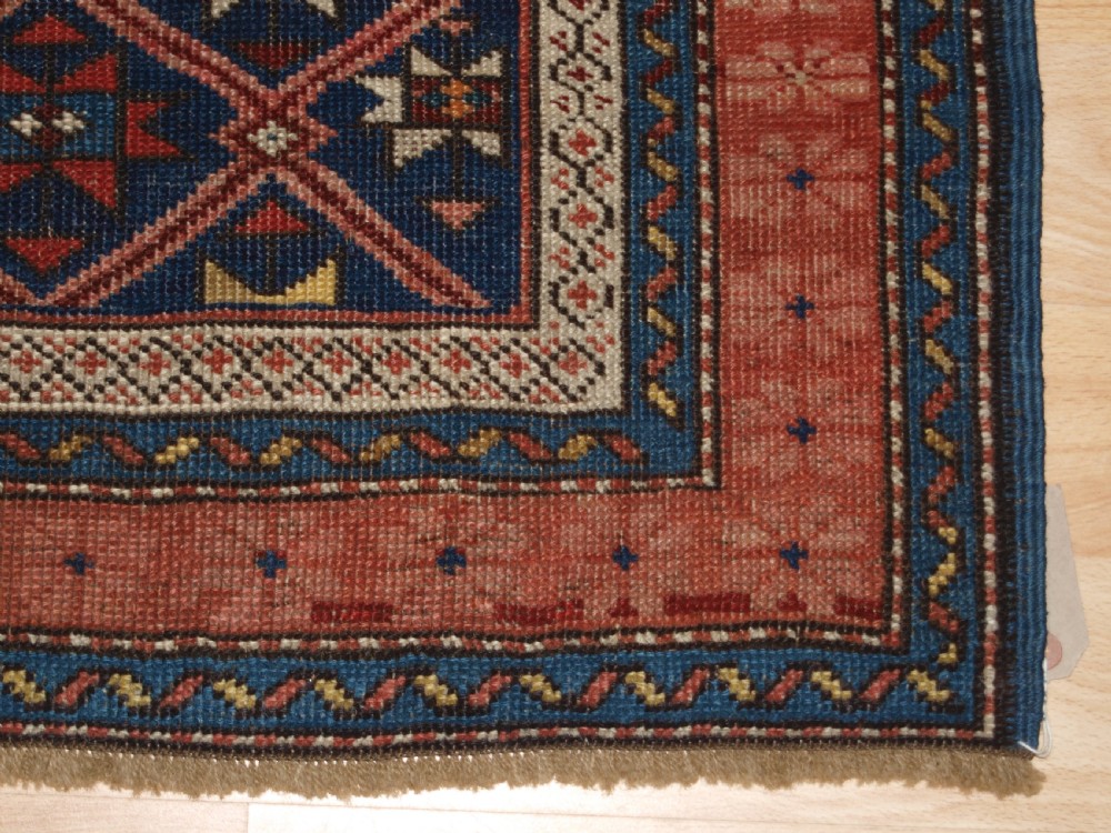 An antique hand-knotted Caucasian rug featuring a central navy blue field with a geometric star-and-lattice pattern framed by a reddish-pink patterned border. View X