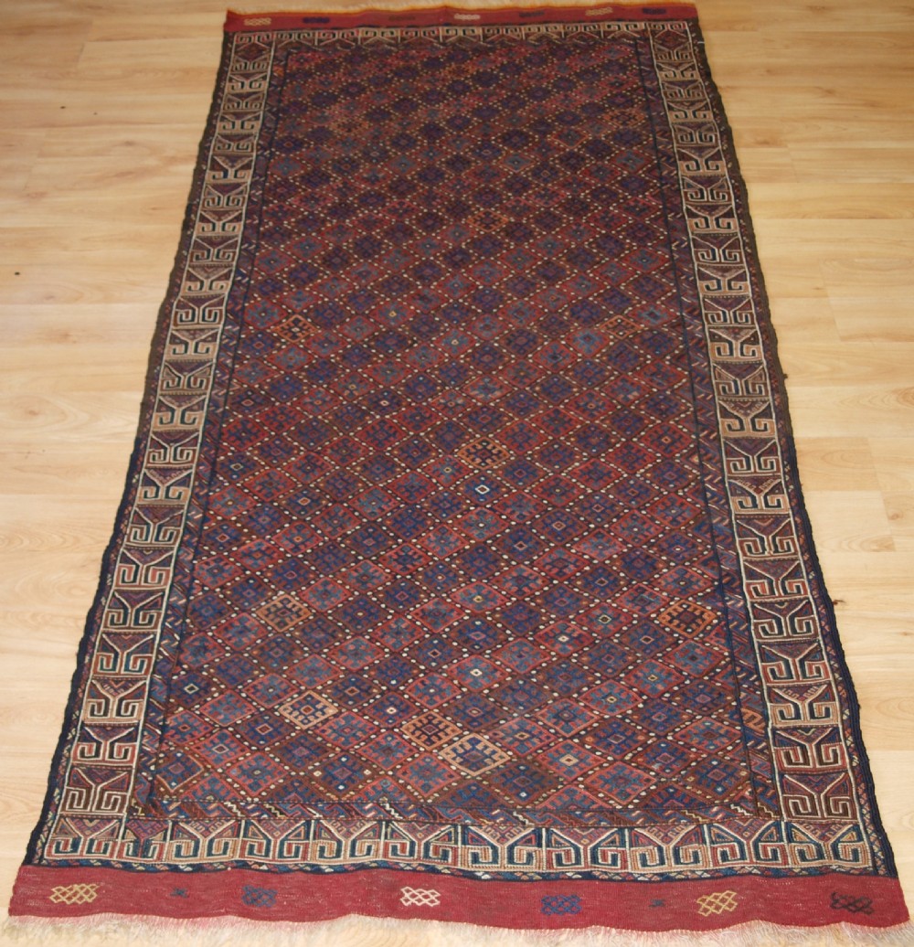 A long handwoven rectangular Kurdish Soumak kilim rug featuring a dense red and blue diamond lattice pattern framed by a geometric meander border.