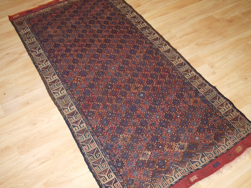 A long handwoven rectangular Kurdish Soumak kilim rug featuring a dense red and blue diamond lattice pattern framed by a geometric meander border. View II